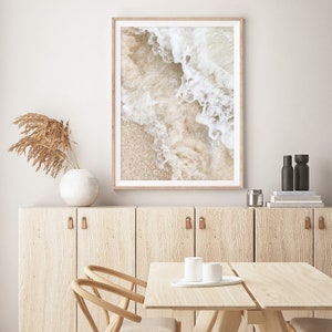 May include: A framed photograph of a beach scene with white foamy waves crashing on the sand. The photo is in a light wood frame and is hanging on a white wall.