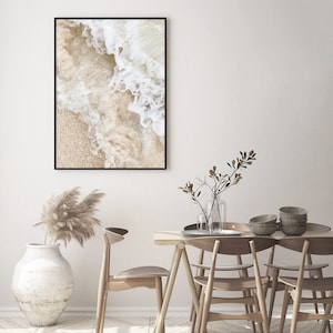 May include: A framed print of a close-up of white ocean waves crashing on a sandy beach. The print is hanging on a white wall above a wooden dining table with chairs.