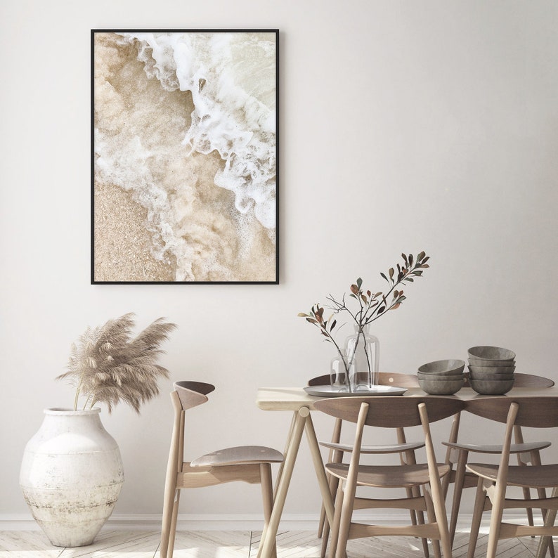 May include: A framed print of a close-up of white ocean waves crashing on a sandy beach. The print is hanging on a white wall above a wooden dining table with chairs.