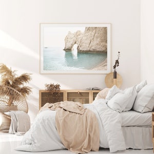 Puede incluir: Una cama blanca con una manta beige y almohadas blancas. Una fotograf&iacute;a enmarcada de un arco rocoso sobre un oc&eacute;ano turquesa cuelga sobre la cama. Una silla de mimbre con un coj&iacute;n de color canela se encuentra en la esquina de la habitaci&oacute;n.