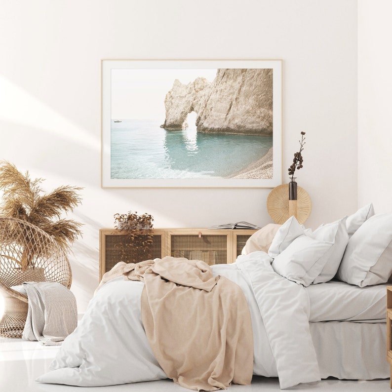 Puede incluir: Una cama blanca con una manta beige y almohadas blancas. Una fotograf&iacute;a enmarcada de un arco rocoso sobre un oc&eacute;ano turquesa cuelga sobre la cama. Una silla de mimbre con un coj&iacute;n de color canela se encuentra en la esquina de la habitaci&oacute;n.