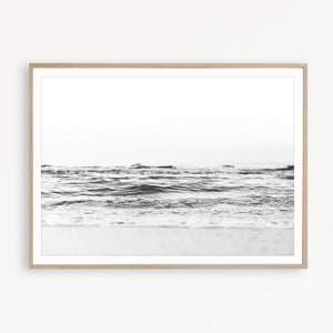 May include: A black and white photograph of ocean waves rolling onto a sandy shore, framed in a light-colored wooden frame. The image captures the low-angle perspective of the water surface with a bright, clear sky visible in the background.