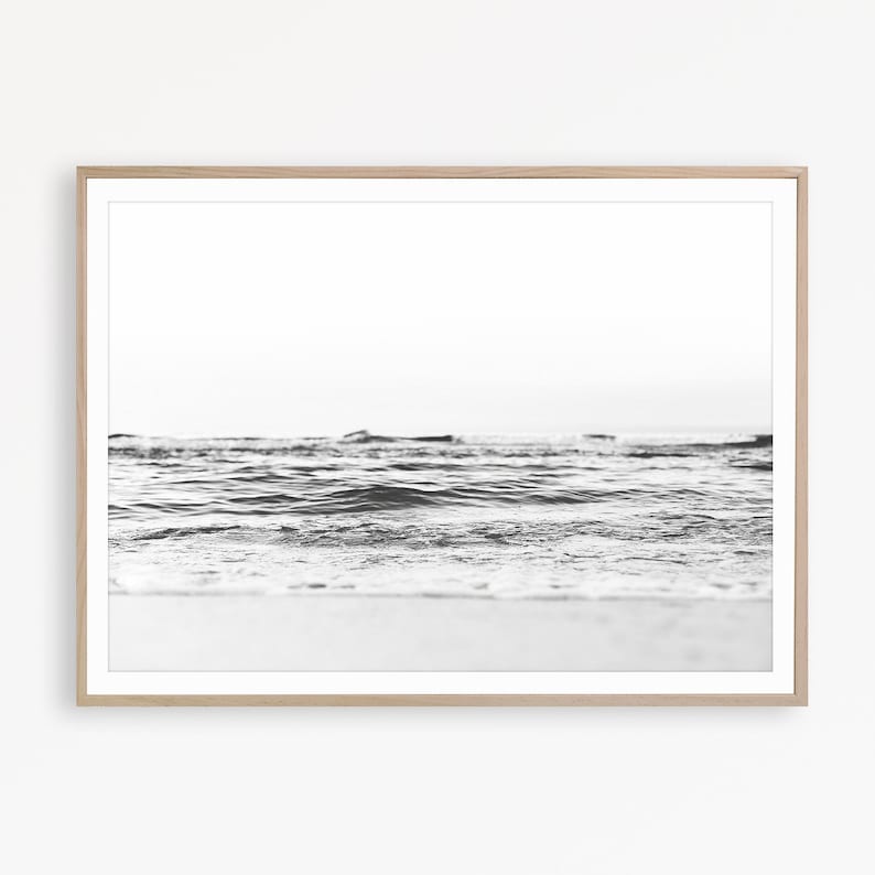 May include: A black and white photograph of ocean waves rolling onto a sandy shore, framed in a light-colored wooden frame. The image captures the low-angle perspective of the water surface with a bright, clear sky visible in the background.