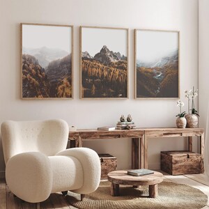 May include: Three vertical framed wall art prints featuring mountain landscapes with golden autumn forests, rocky peaks, and misty valleys. The artwork is displayed in light wood frames, showcasing a consistent color palette of brown, gold, and grey.