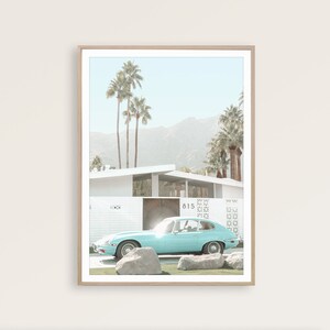 May include: A light blue vintage car parked in front of a white mid-century modern house with palm trees and mountains in the background. The house has a number "815" on the side.