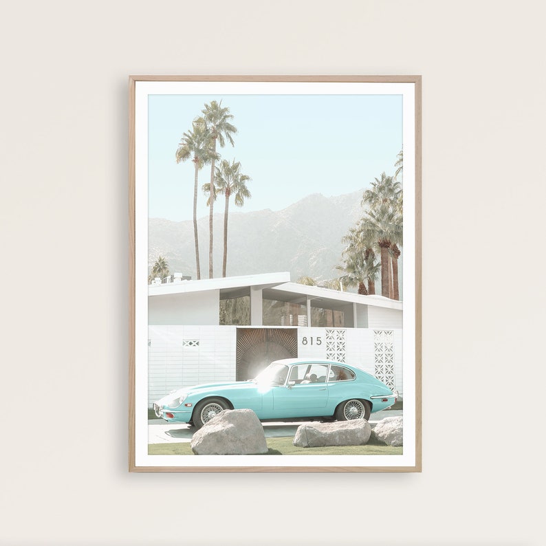 May include: A light blue vintage car parked in front of a white mid-century modern house with palm trees and mountains in the background. The house has a number "815" on the side.
