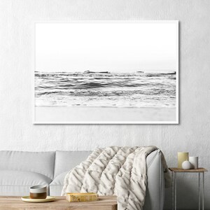 May include: A black and white framed wall art print depicting a low-angle view of ocean waves rolling onto a sandy beach. The horizon line is visible in the distance, with the water showing textured ripples and foam against the shoreline.
