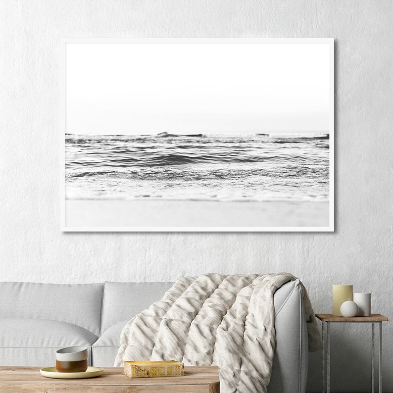 May include: A black and white framed wall art print depicting a low-angle view of ocean waves rolling onto a sandy beach. The horizon line is visible in the distance, with the water showing textured ripples and foam against the shoreline.