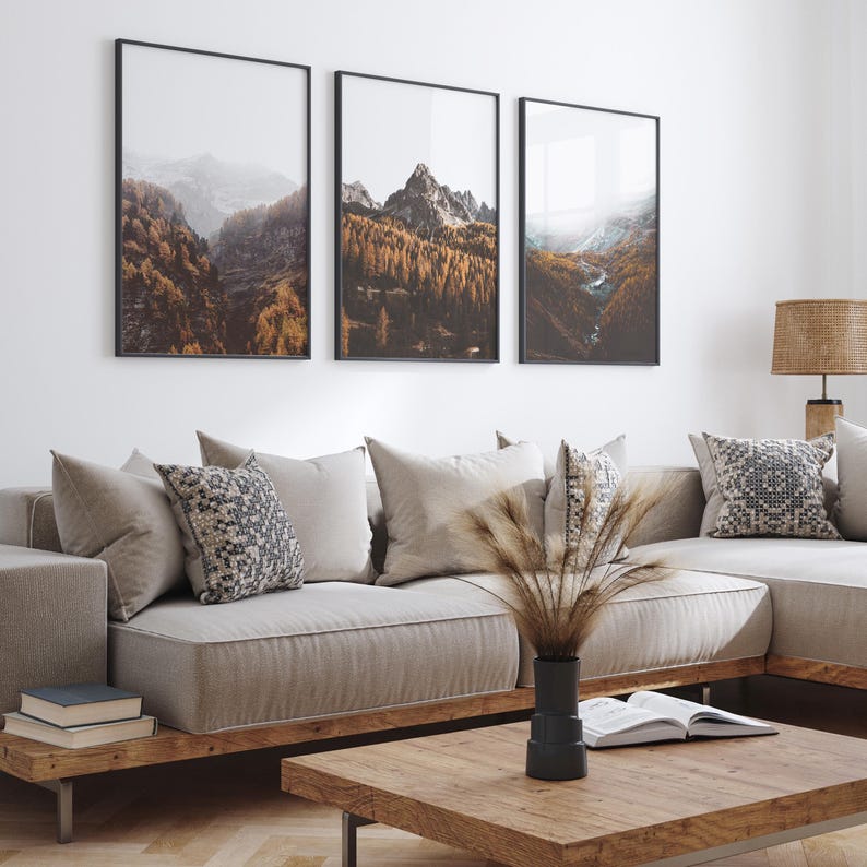 May include: A triptych of framed wall art prints featuring mountain landscapes with golden autumn forests and misty peaks, displayed above a beige sectional sofa with patterned throw pillows and a wooden coffee table with a black vase.