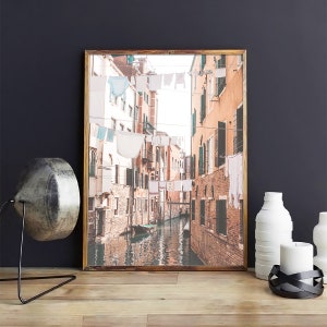 May include: A photograph of a narrow canal in Venice, Italy.  The canal is lined with buildings and there are clothes hanging on lines strung across the canal.  A small boat is in the water.