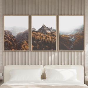 May include: A triptych of three vertical framed wall art prints depicting a mountain landscape. The images feature jagged rocky peaks, dense forests with golden-brown autumn foliage, and misty valleys under a bright, overcast sky.