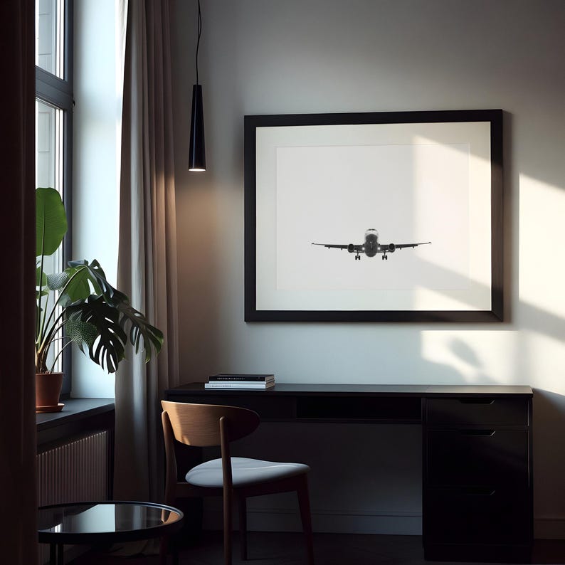 May include: A monochrome framed print of an aeroplane in flight is displayed above a black desk. A wooden chair with a grey seat is in front of the desk. A potted plant sits near a window with a sheer curtain.