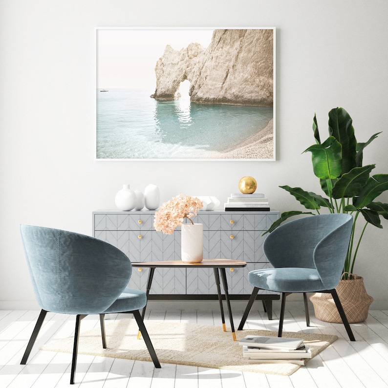 Puede incluir: Un dise&ntilde;o de interior de sala de estar con dos sillas azules, una mesa de caf&eacute;, un aparador y una fotograf&iacute;a enmarcada de una playa rocosa con un arco natural. El aparador tiene un acabado gris claro con un patr&oacute;n de chevron y pomos dorados. La mesa de caf&eacute; est&aacute; hecha de madera con un acabado marr&oacute;n oscuro y patas de metal negro. Las sillas est&aacute;n tapizadas en una tela azul suave y tienen patas de metal negro. La fotograf&iacute;a est&aacute; en un marco blanco y presenta un paisaje mar&iacute;timo con un arco rocoso. La habitaci&oacute;n est&aacute; decorada con una planta en maceta y un jarr&oacute;n de flores rosas.