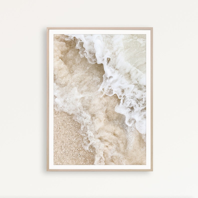 May include: A close-up photograph of white ocean waves crashing on a sandy beach. The sand is a light beige color and the waves are foamy and white.