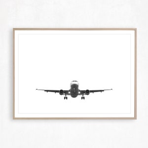 May include: A monochrome photograph of an aeroplane in flight, centrally positioned. The plane is viewed head-on, with its wings spread. The image is framed in a light-coloured wooden frame, set against a white background.