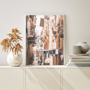 May include: A photograph of a narrow street in a city with buildings on both sides. The buildings are painted in shades of beige and brown. Laundry is hanging on lines strung across the street.
