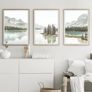 May include: Three framed landscape photographs of a lake with mountains and trees in the background. The lake is a light blue color and the mountains are a light gray color. The trees are a dark green color. The frames are a light brown color.