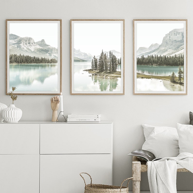 May include: Three framed landscape photographs of a lake with mountains and trees in the background. The lake is a light blue color and the mountains are a light gray color. The trees are a dark green color. The frames are a light brown color.