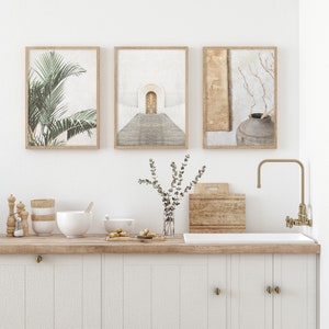 May include: Three framed prints with minimalist designs. The first print features a green palm leaf, the second print features a white archway with a wooden door, and the third print features a brown vase with a dried branch.
