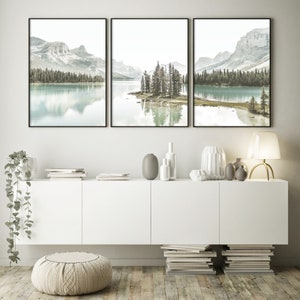 May include: A set of three framed landscape photographs featuring a lake with mountains in the background. The lake is a light blue color and the mountains are a light gray color. The trees are a dark green color. The frames are black.