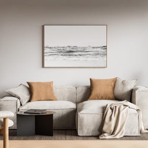 May include: A light gray modular sofa with two tan square throw pillows and a draped beige blanket. A black circular side table sits to the left, and a framed black and white landscape print of the ocean hangs on the wall above the sofa.