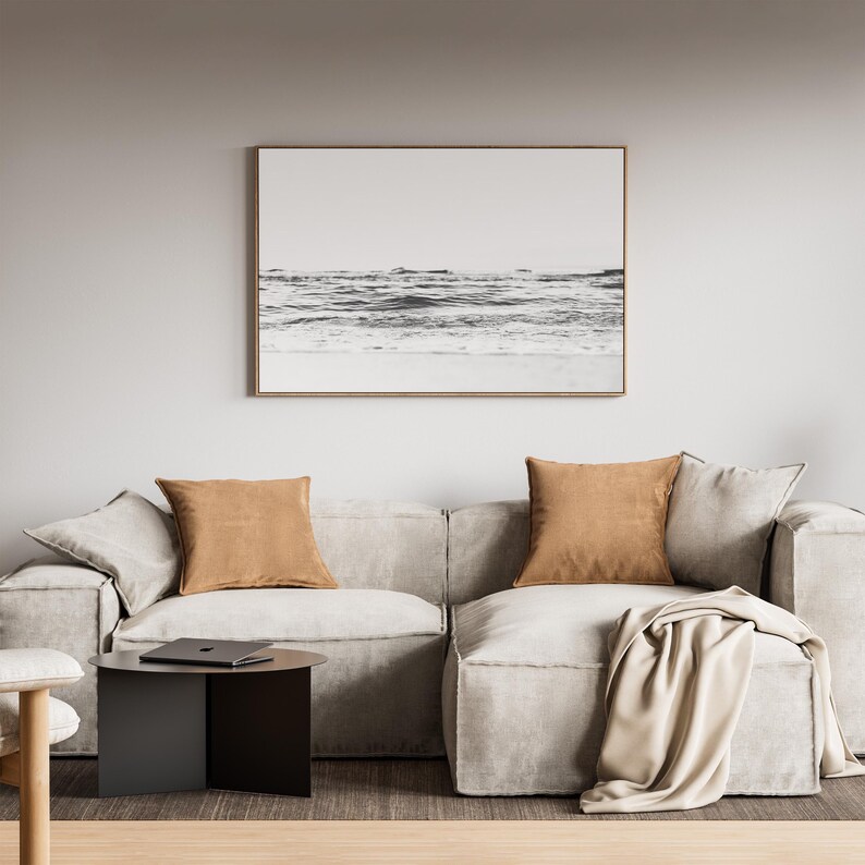 May include: A light gray modular sofa with two tan square throw pillows and a draped beige blanket. A black circular side table sits to the left, and a framed black and white landscape print of the ocean hangs on the wall above the sofa.