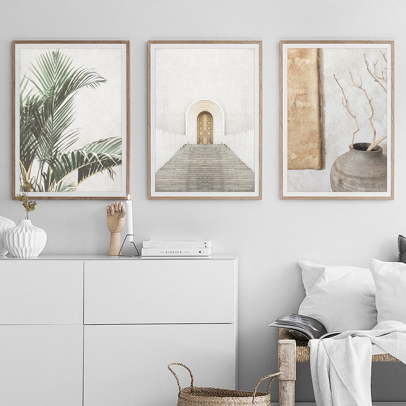 May include: Three framed prints with minimalist designs. The first print features a green palm leaf, the second print shows a white archway with a wooden door and steps leading up to it, and the third print shows a brown vase with a branch of twigs.