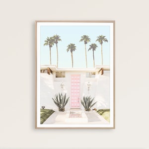 May include: A white mid-century modern house with a bright pink door and palm trees in the background. The house has a white exterior and a concrete walkway leading to the front door.