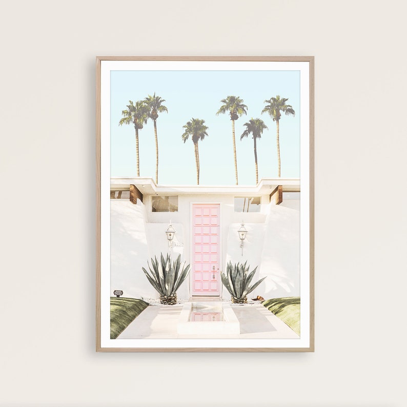 May include: A white mid-century modern house with a bright pink door and palm trees in the background. The house has a white exterior and a concrete walkway leading to the front door.
