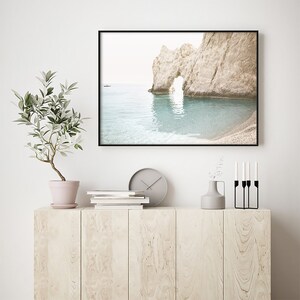 Puede incluir: Una fotograf&iacute;a enmarcada de un arco rocoso en una playa con un peque&ntilde;o barco en la distancia. El agua es de color azul claro y el cielo es de un blanco brumoso. La foto est&aacute; colgada en una pared blanca sobre un armario de madera con un reloj, libros, un jarr&oacute;n y candelabros.