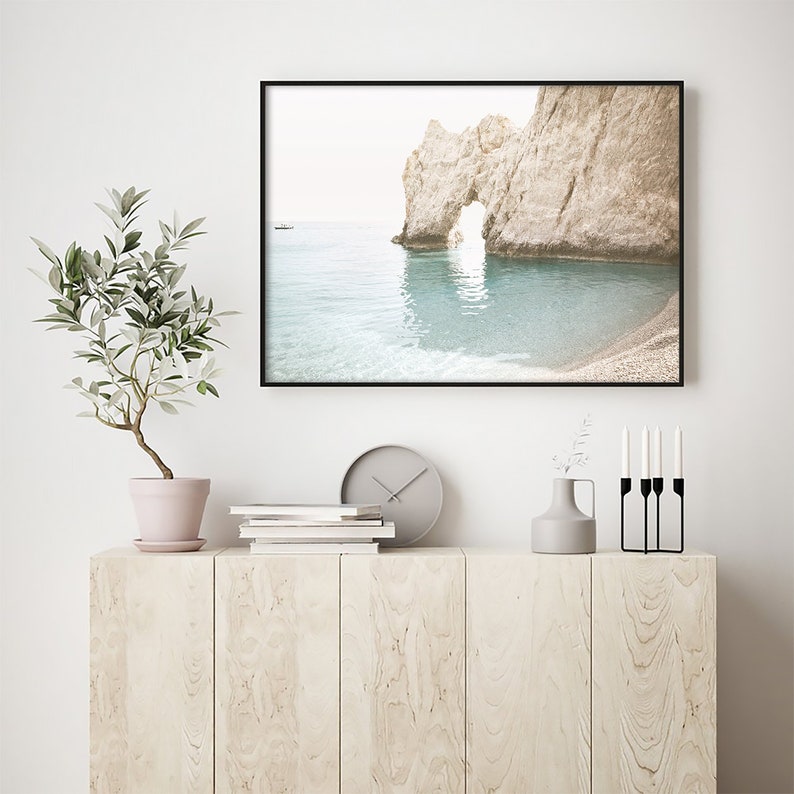 Puede incluir: Una fotograf&iacute;a enmarcada de un arco rocoso en una playa con un peque&ntilde;o barco en la distancia. El agua es de color azul claro y el cielo es de un blanco brumoso. La foto est&aacute; colgada en una pared blanca sobre un armario de madera con un reloj, libros, un jarr&oacute;n y candelabros.