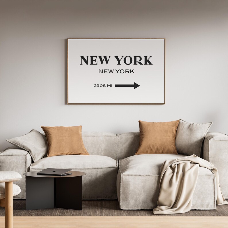 May include: A rectangular wall art print in a thin frame featuring the text NEW YORK, NEW YORK and 2908 MI with a black arrow pointing to the right. The print has a white background with bold black lettering and is displayed on a wall.