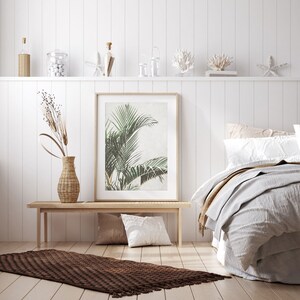 May include: A white bedroom with a wooden bench, a brown woven rug, and a framed print of a tropical plant. The bed has a white duvet and a brown throw blanket.