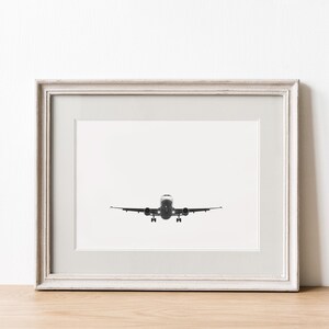 May include: Framed black and white photograph of an airplane in flight, viewed head-on. The plane is centered against a white background, with its landing gear extended. The photograph is in a white distressed wooden frame.