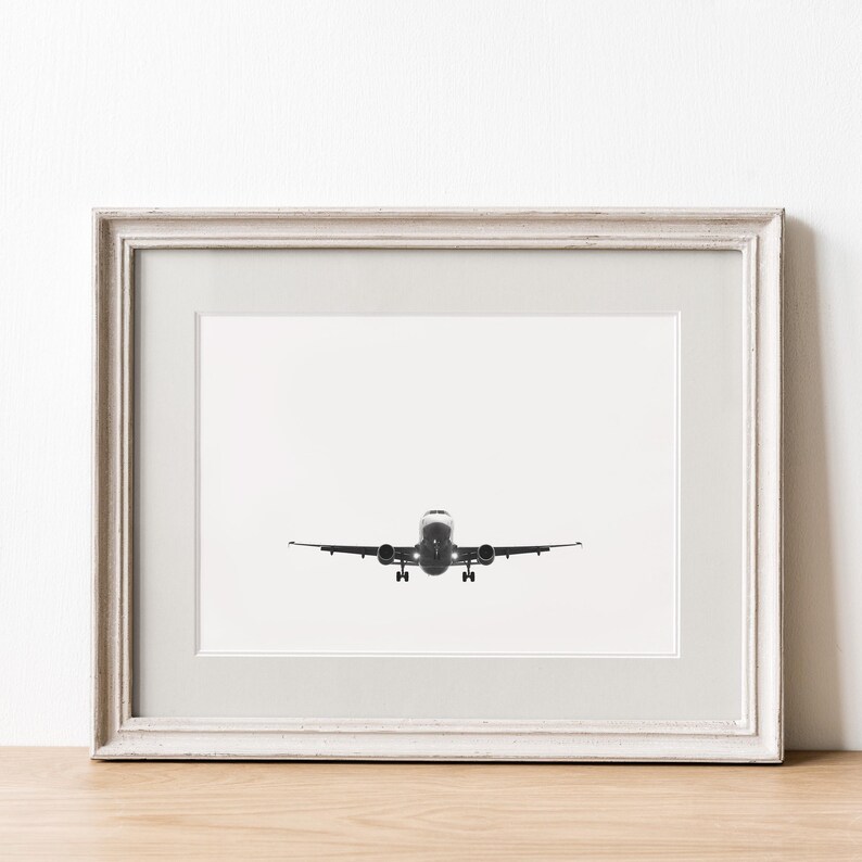 May include: Framed black and white photograph of an airplane in flight, viewed head-on. The plane is centered against a white background, with its landing gear extended. The photograph is in a white distressed wooden frame.