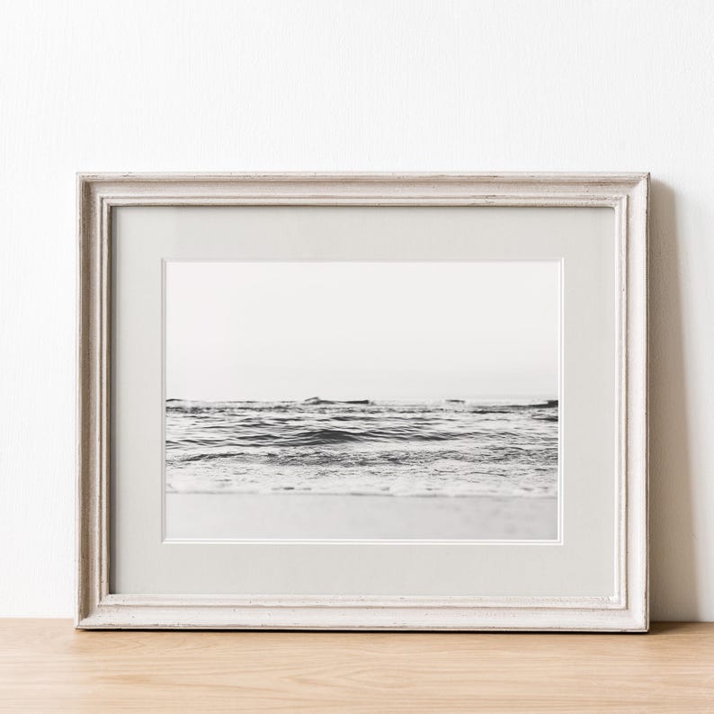 May include: A black and white photograph of ocean waves meeting the shoreline, displayed in a light-colored wooden frame with a wide white mat. The image captures the horizon line and the gentle movement of the water in a horizontal orientation.