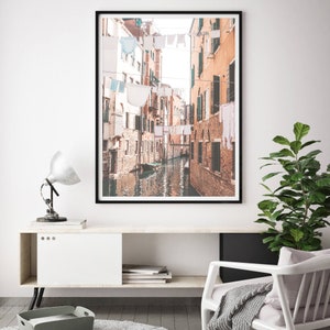 May include: A black and white photograph of a narrow canal in Venice, Italy. Buildings line the canal, with laundry hanging from lines strung across the water. A small boat is visible in the canal.