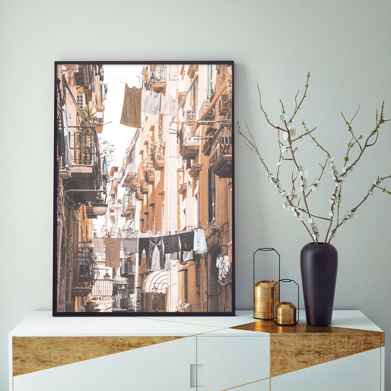 May include: A framed photograph of a narrow, sunlit street in a European city. The buildings are painted in warm, earthy tones, and laundry hangs on lines strung across the street.