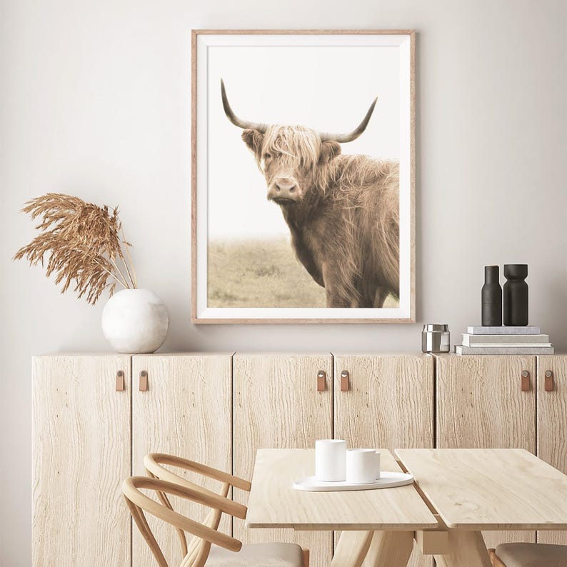 Może przedstawiać: Oprawiony wydruk przedstawiający szkocką krowę rasy Highland, z długim, brązowym futrem i dużymi rogami. Grafika umieszczona na miękkim tle, tworząc rustykalny nastr&oacute;j. Wydruk w jasnej drewnianej ramie, idealny do dekoracji.