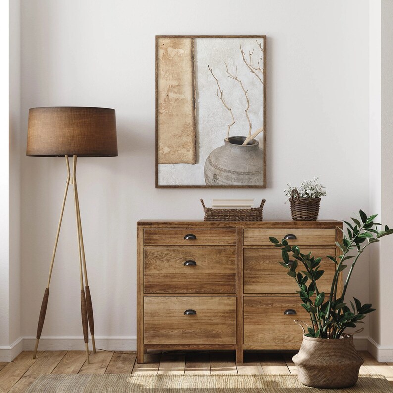 May include: A wooden dresser with four drawers and black handles. A brown lamp with a brown shade sits on the left side of the dresser. A potted plant with green leaves sits on the right side of the dresser. A framed print of a vase with branches hangs on the wall above the dresser.