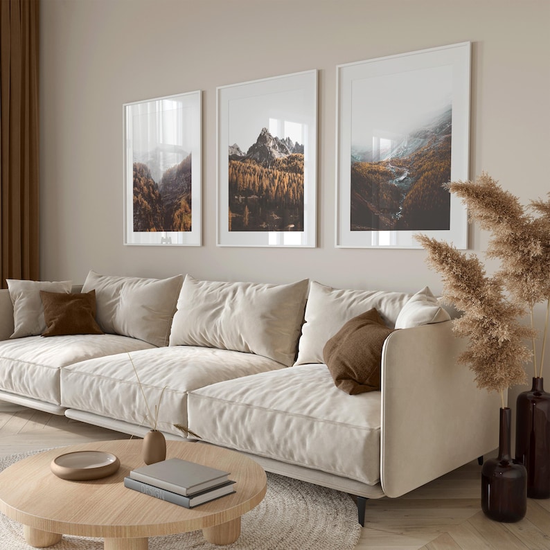 May include: Three vertical framed wall art prints featuring mountain landscapes with autumn forests and rocky peaks, displayed in white frames above a beige sofa with brown accent pillows and a round wooden coffee table with books.