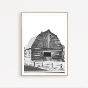 May include: Black and white photograph of a wooden barn with a large double door and a smaller window. The barn is surrounded by a wooden fence and a dirt road.