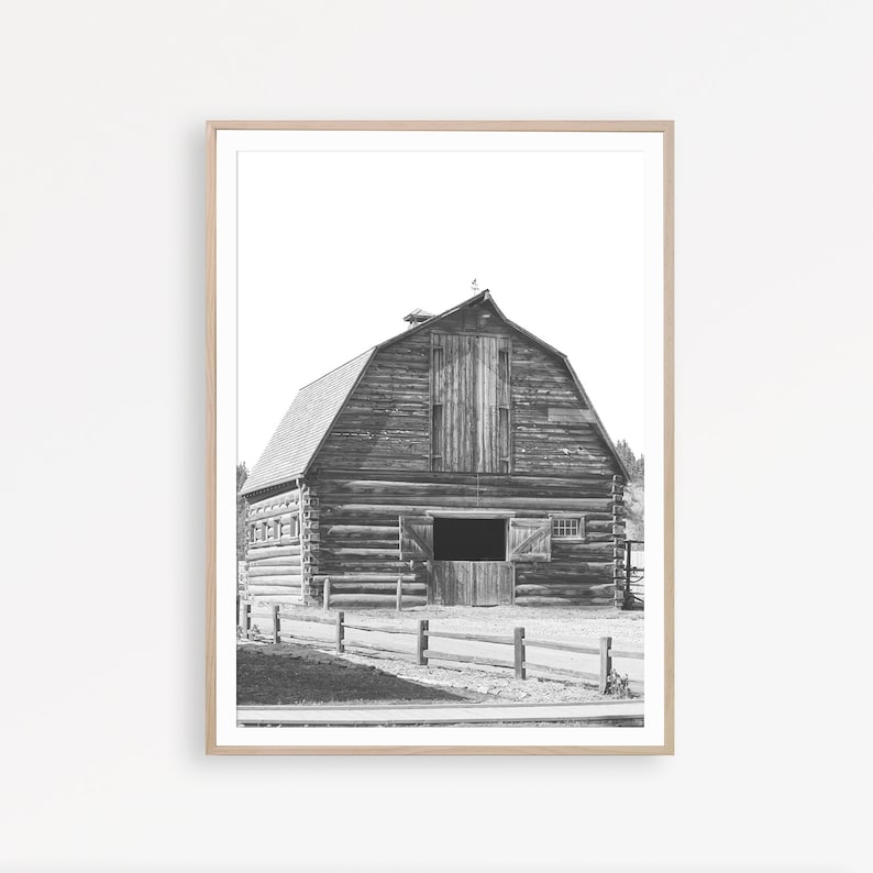 May include: Black and white photograph of a wooden barn with a large double door and a smaller window. The barn is surrounded by a wooden fence and a dirt road.