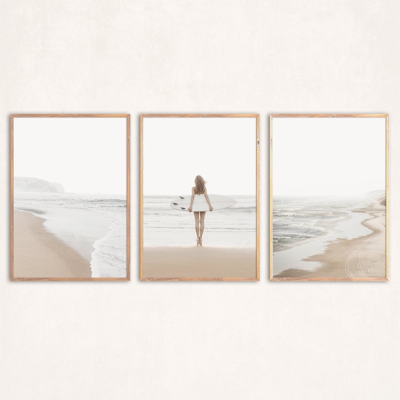 May include: A set of three framed prints featuring a woman standing on a beach with a surfboard. The prints are in a minimalist style with a white background and a light brown frame.