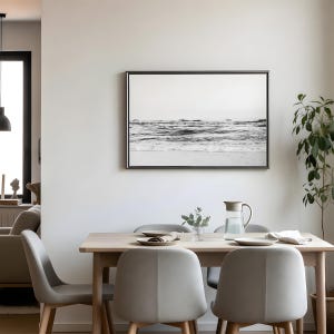 May include: A rectangular wall art piece featuring a black and white photograph of ocean waves crashing against a shoreline, enclosed in a thin dark frame, displayed above a light wood dining table with four gray chairs and a glass water pitcher.