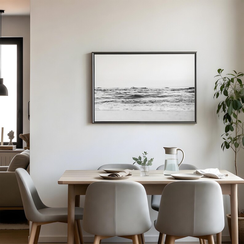 May include: A rectangular wall art piece featuring a black and white photograph of ocean waves crashing against a shoreline, enclosed in a thin dark frame, displayed above a light wood dining table with four gray chairs and a glass water pitcher.