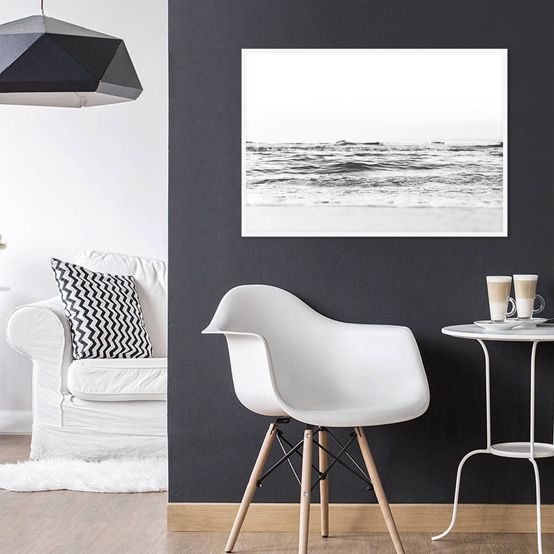 May include: A black and white framed landscape photograph of ocean waves hanging on a dark gray wall above a white molded plastic chair with wooden legs, positioned near a white side table with two glass mugs and a white sofa with a patterned pillow.