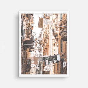 May include: A narrow, sun-drenched alleyway in a European city with clothes hanging on lines strung between buildings. The buildings are a warm, pale yellow with balconies and windows.