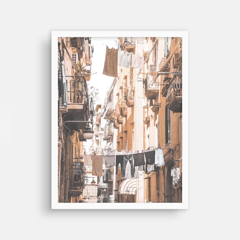 May include: A narrow, sun-drenched alleyway in a European city with clothes hanging on lines strung between buildings. The buildings are a warm, pale yellow with balconies and windows.