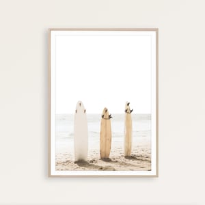 Puede incluir: Tres tablas de surf est&aacute;n de pie en una playa de arena. Las tablas de surf son de diferentes colores, una blanca, otra marr&oacute;n y otra beige. Las tablas de surf est&aacute;n todas apoyadas la una en la otra, y la playa est&aacute; vac&iacute;a.